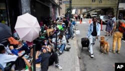 FILE - Migrants sit in a queue outside of the Roosevelt Hotel, which is being used by the city as temporary housing for migrants bused to New York from Texas, July 31.