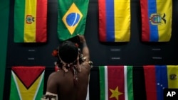 Seorang pria pribumi mengulurkan tangannya di atas bendera Brazil di paviliun delapan negara Amerika Selatan yang berbagi wilayah hutan hujan Amazon, selama pertemuan Dialog Amazon di di Belem, Brazil, 6 Agustus 2023. (Foto: AP)