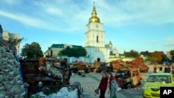 Two women holding bouquets of flowers stand in front of a display of destroyed Russian tanks and armored vehicles near St. Michael's Golden-Domed Monastery in Kyiv, Sept. 11, 2022