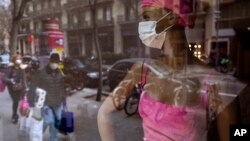 Warga yang membawa persediaan bahan pokok mengenakan masker, berjalan di depan manekin yang terpajang di sebuah toko di pusat kota Barcelona, Spanyol, 14 Maret 2020.