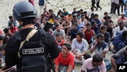 Seorang polisi berjaga-jaga saat pria etnis Rohingya duduk di pantai seusai mendarat di Aceh Besar, Minggu, 8 Januari 2023. Lebih dari 100 pengungsi Muslim Rohingya tiba di Aceh setelah perjalanan panjang dan berbahaya di atas perahu kayu. (AP/Muhammad Ahyar)