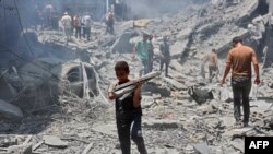 Seorang anak laki-laki membawa barang-barang yang berhasil diselamatkan dari puing-puing bangunan yang hancur akibat pengeboman Israel di kamp pengungsi al-Shati di Kota Gaza pada 22 Juni 2024. (Foto: AFP)