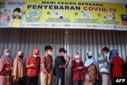 Warga berdiri di bawah spanduk yang mengingatkan warga untuk memakai masker dan tips lainnya dalam memerangi Covid-19 saat mereka mengantri untuk membeli minyak goreng di Bandung pada 18 Februari 2022. (Foto: AFP)
