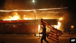 បាតុករម្នាក់កាន់ទង់ជាតិសហរដ្ឋអាមេរិកឡើងលើដែលជាសញ្ញាមួយនៃមិនពេញចិត្ត នៅក្បែរអាគារដែលកំពុងឆេះកាលពីថ្ងៃព្រហស្បតិ៍ទី ២៨ ខែឧសភាឆ្នាំ ២០២០ នៅទីក្រុង Minneapolis។ (AP / Julio Cortez) 