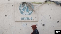 Seorang perempuan Palestina berjalan melewati tembok rusak yang bertuliskan logo UNRWA di kamp pengungsi internal di Rafah, Jalur Gaza selatan, 28 Mei 2024. (Foto: AFP)